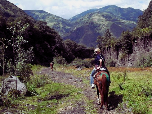 A white woman with blonde hair in casual dress looks over her shoulder at the camera. She is riding a horse. Another horse is farther off down the trail. The backdrop is of the Andes in Ecuador.